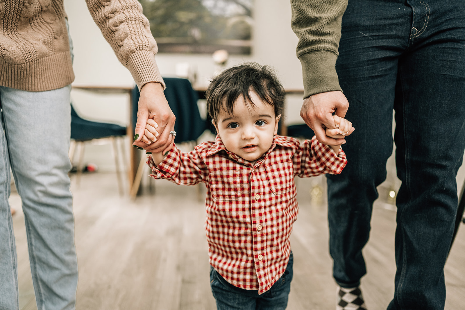 Toddler taking first steps while holding parents’ hands, documenting the first year of newborn photos.