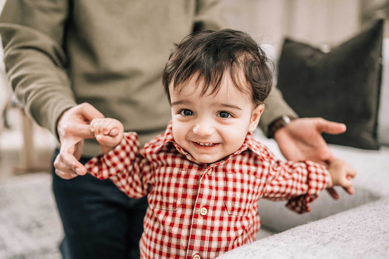 Smiling toddler in red gingham shirt taking first steps, supported by an adult’s hands in a cozy home setting.