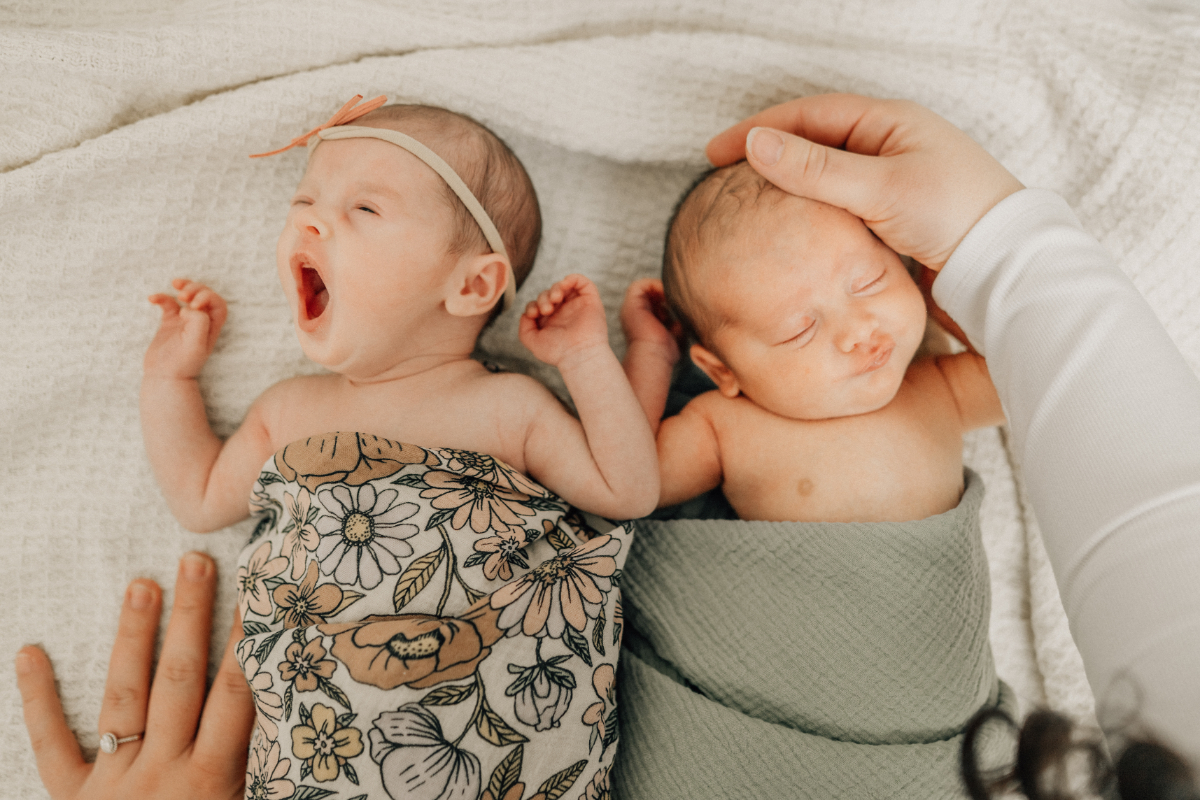 Twin newborns lying next to each other on a bed, one yawning while a parent gently rests a hand nearby.