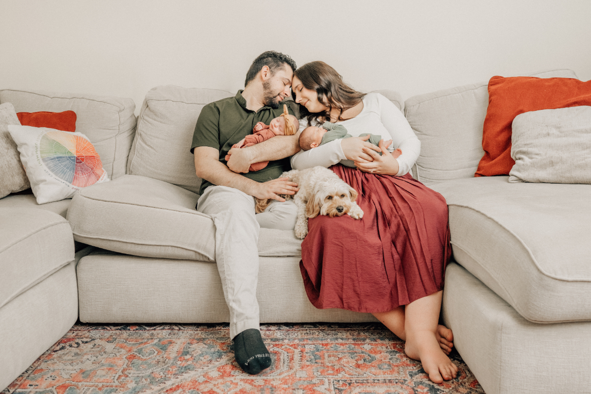 Parents sitting together on a couch holding their newborn while their dog rests nearby during an in-home newborn photography session.
