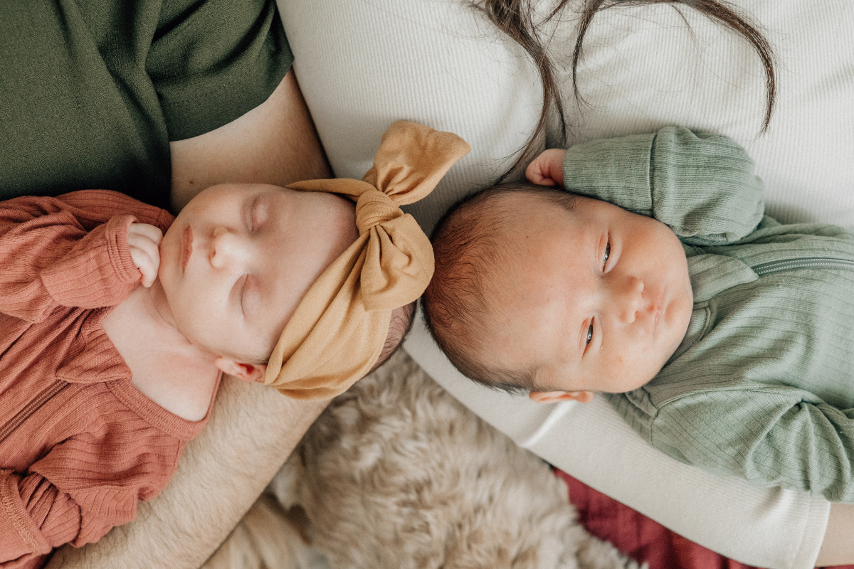 Twin newborns resting side by side in a parent’s arms, dressed in soft neutral outfits during an in-home newborn photography session.