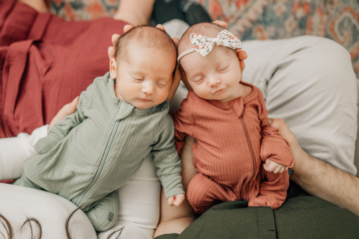 Twin newborns sleeping side by side in soft outfits, resting closely together on a neutral surface.
