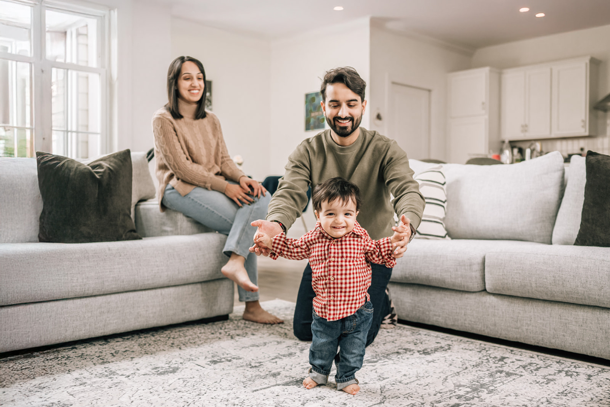 Parents helping their toddler take early steps in the living room while another parent watches nearby.