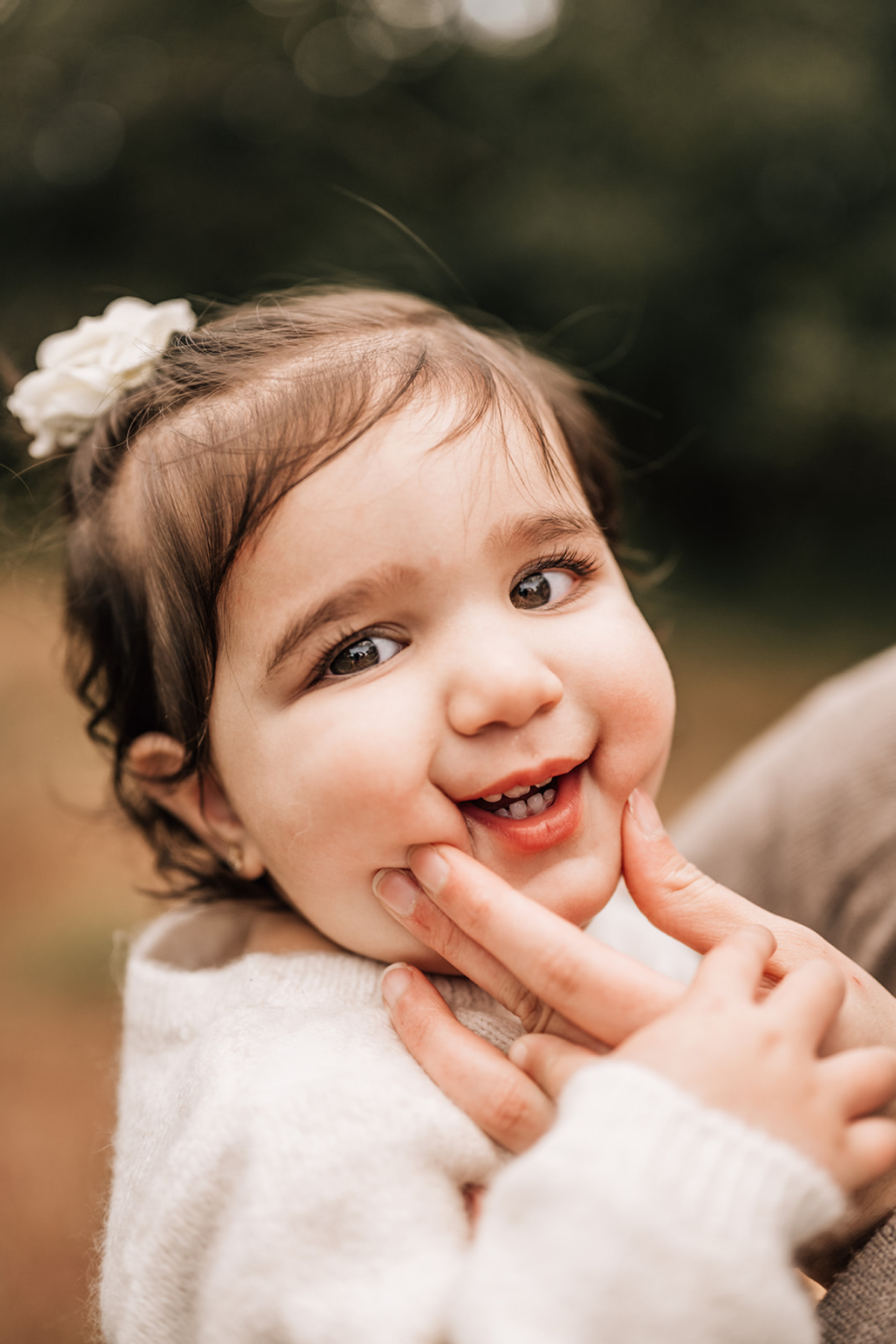 Close-up of smiling baby outdoors as a parent gently touches their cheeks during a playful moment.