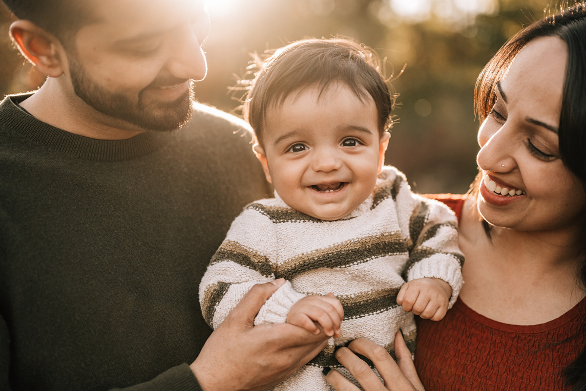 Parents holding smiling baby outdoors in warm light, sharing a relaxed moment together as a family.