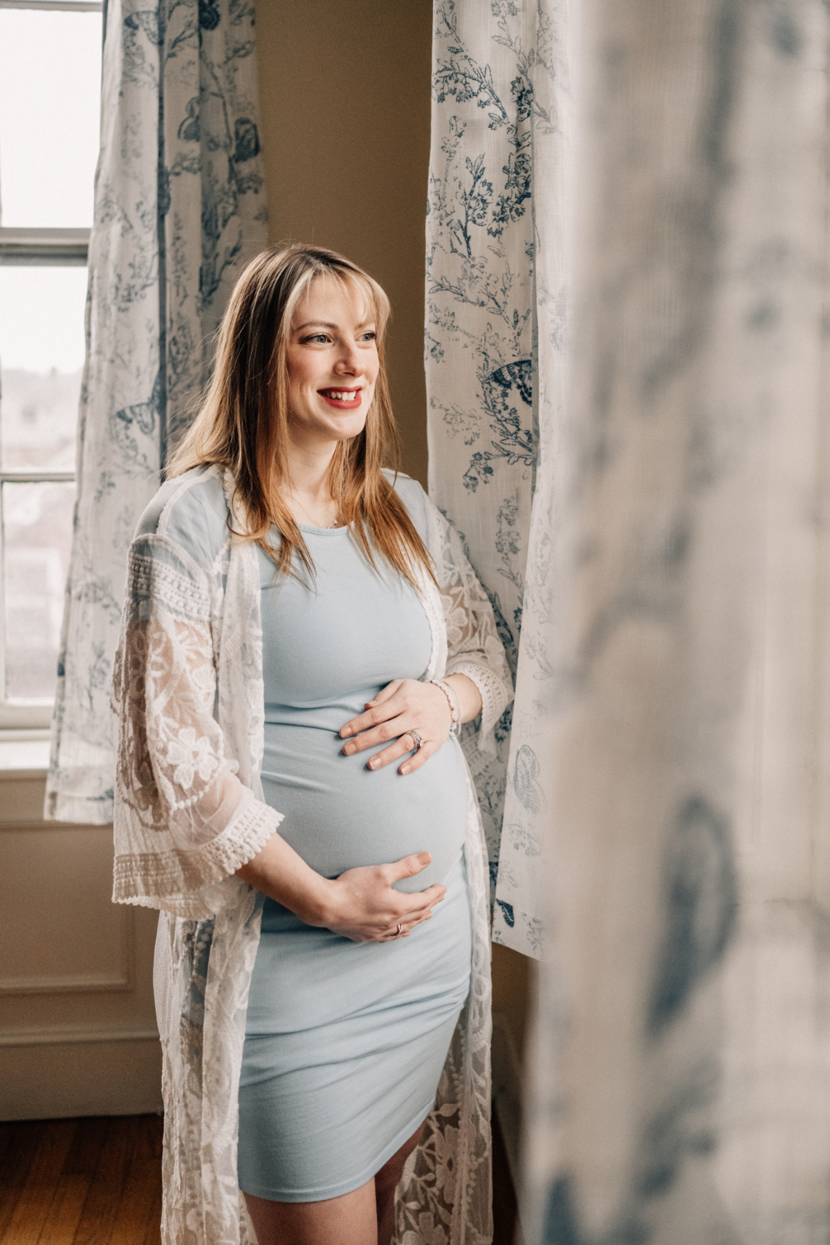 Expecting mother standing by a window indoors during milestone photography session, capturing quiet anticipation and connection.