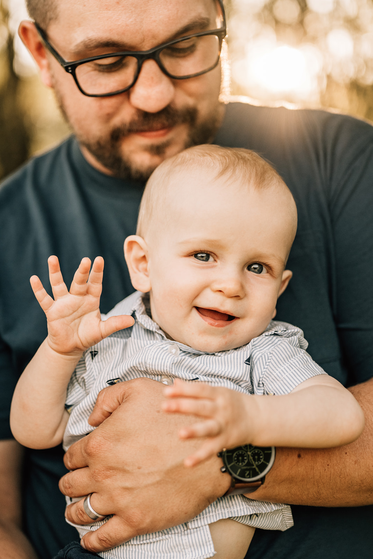 Parent holding smiling baby outdoors, capturing a playful moment as the baby waves and looks toward the camera.