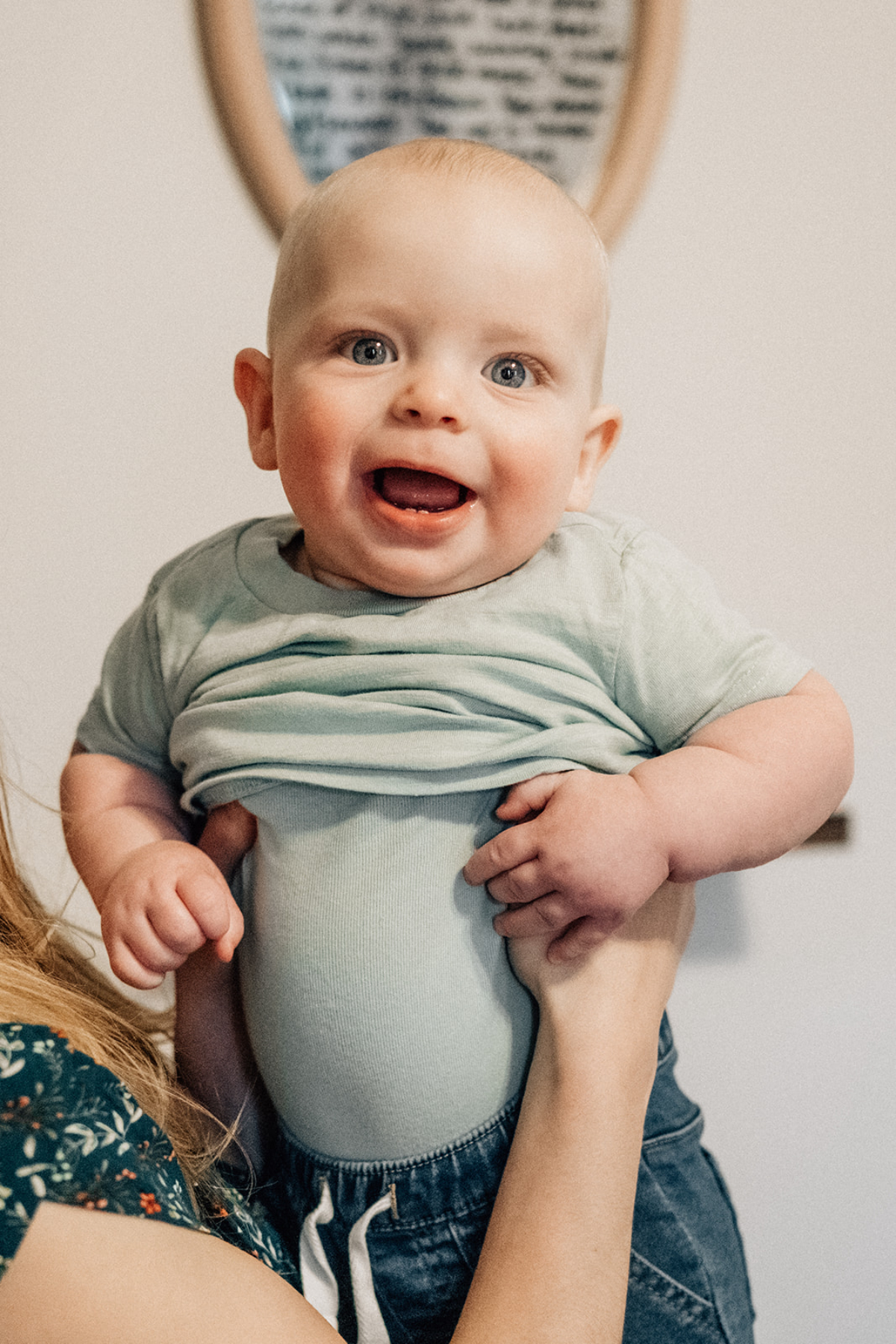 Smiling baby being supported indoors, showing bright eyes and a joyful expression during a relaxed moment at home.