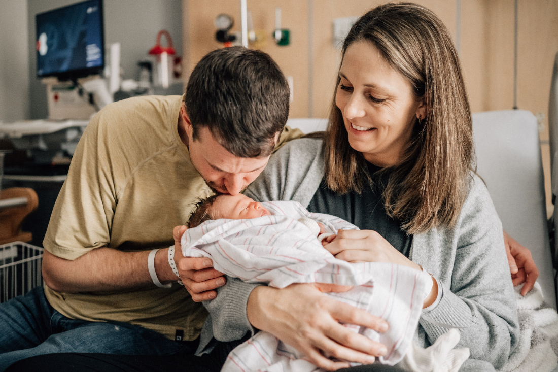 Father kissing his newborn’s forehead as both parents sit together on a hospital bed.