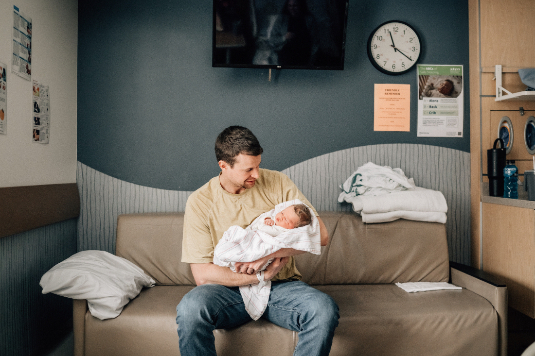 Father sitting on a hospital couch holding his newborn, documenting calm connection during fresh 48 photos.