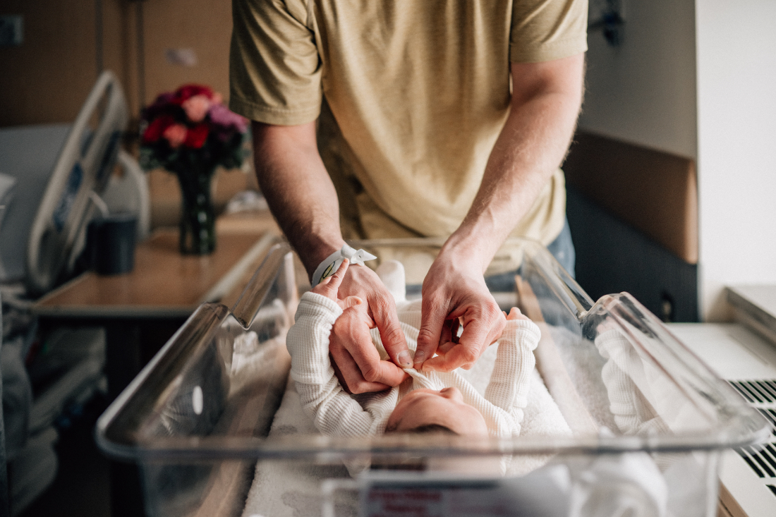 Parent gently fastening newborn clothing while baby lies in a clear hospital bassinet.