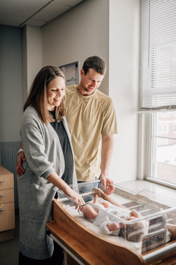 Parents standing together beside a hospital bassinet, looking down at their newborn during the first day together.