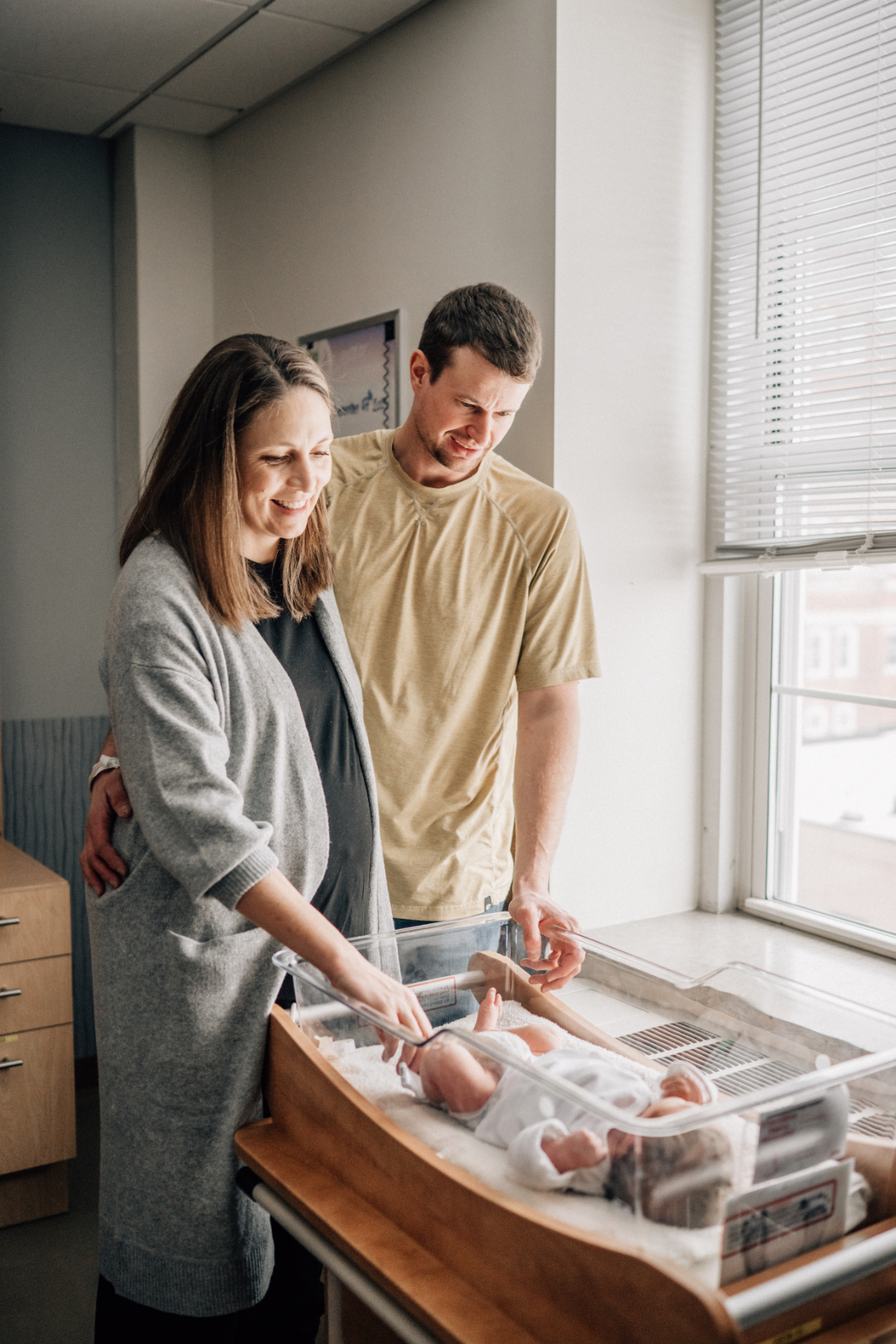 Parents standing together beside a hospital bassinet, looking down at their newborn during the first day together.