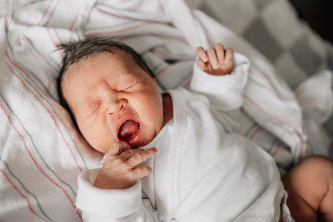 Newborn yawning peacefully in a hospital bassinet, wrapped in a white outfit and striped blanket.