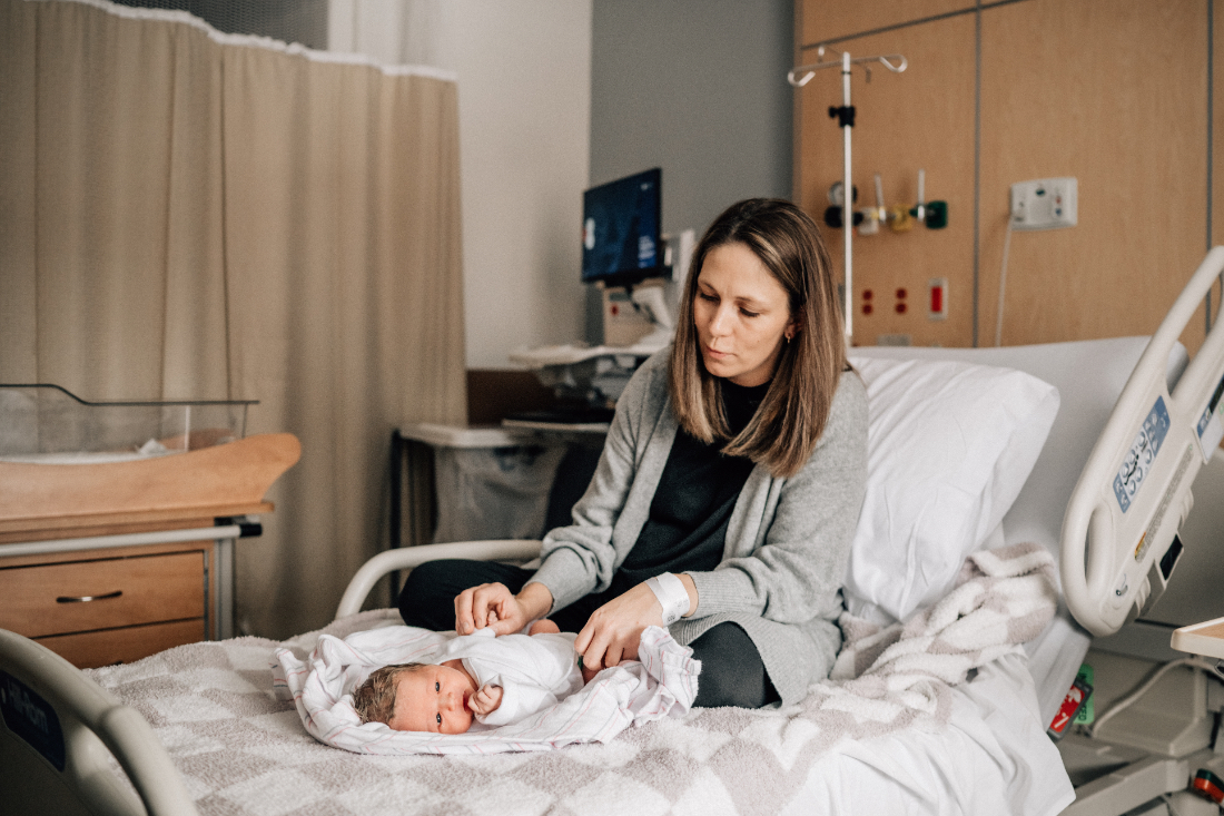 Mother sitting on a hospital bed, gently adjusting her newborn during a calm fresh 48 photo session.
