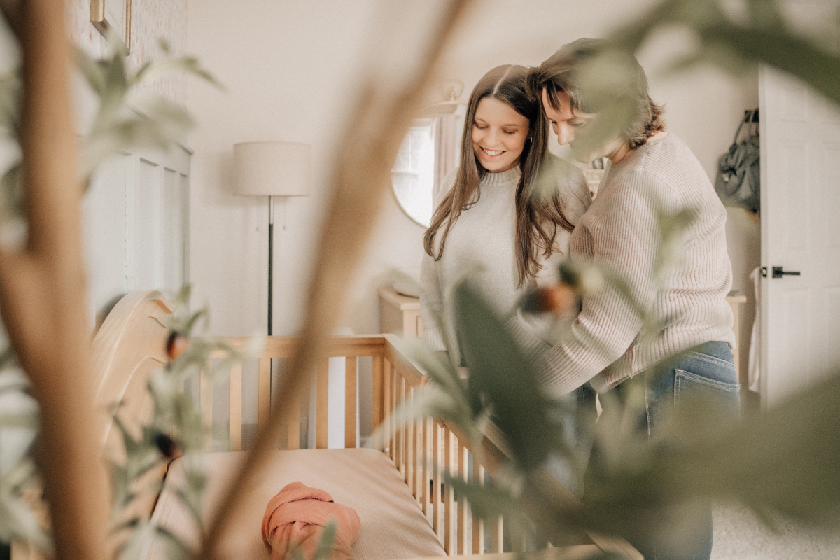 Two moms standing by crib, smiling down at sleeping baby in soft nursery light