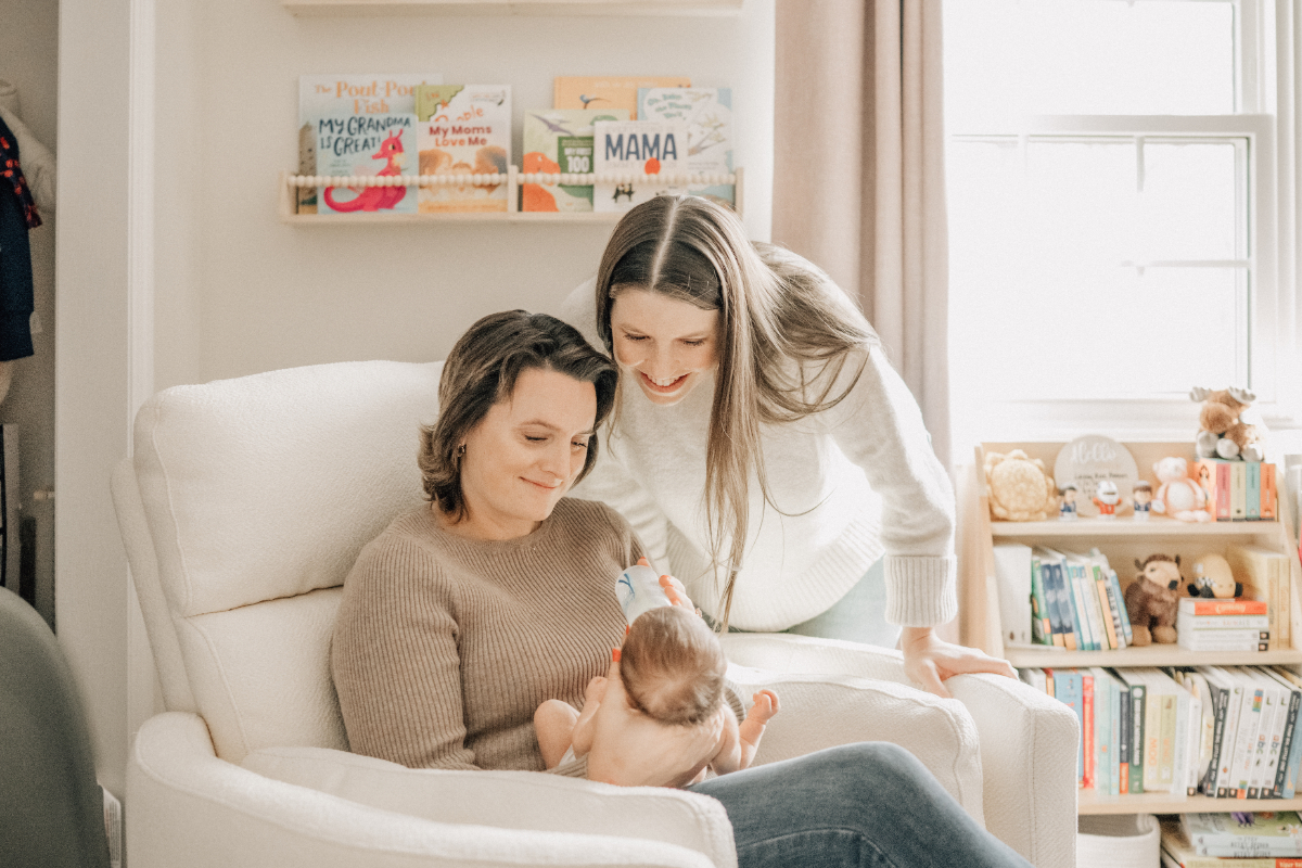 Two moms smiling at baby during quiet moment in nursery chair.