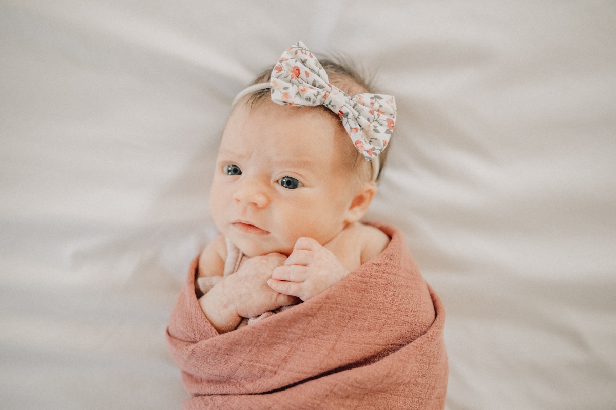 Baby girl wrapped in rust swaddle, wearing floral bow headband, wide awake on bed