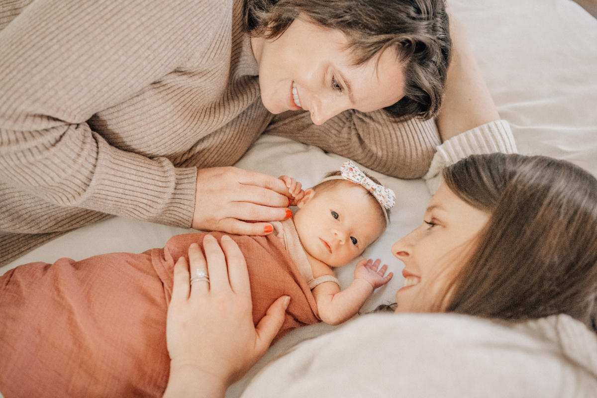 Two moms lying beside baby girl on bed, smiling and gently touching her face