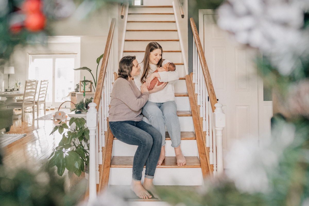 Two moms sitting on staircase, admiring newborn in soft home setting.
