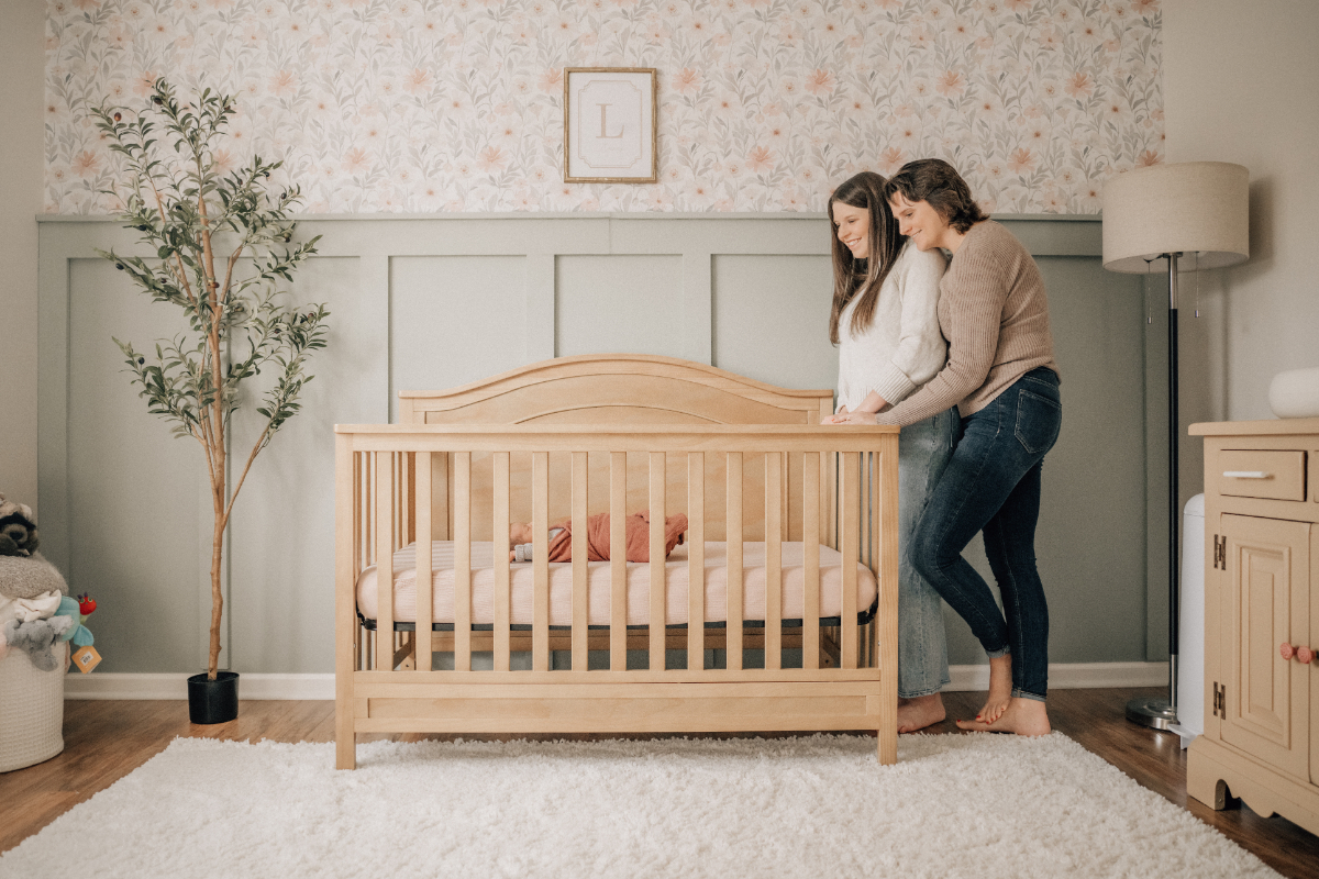 In-home newborn baby photos of two moms standing by crib in soft nursery.
