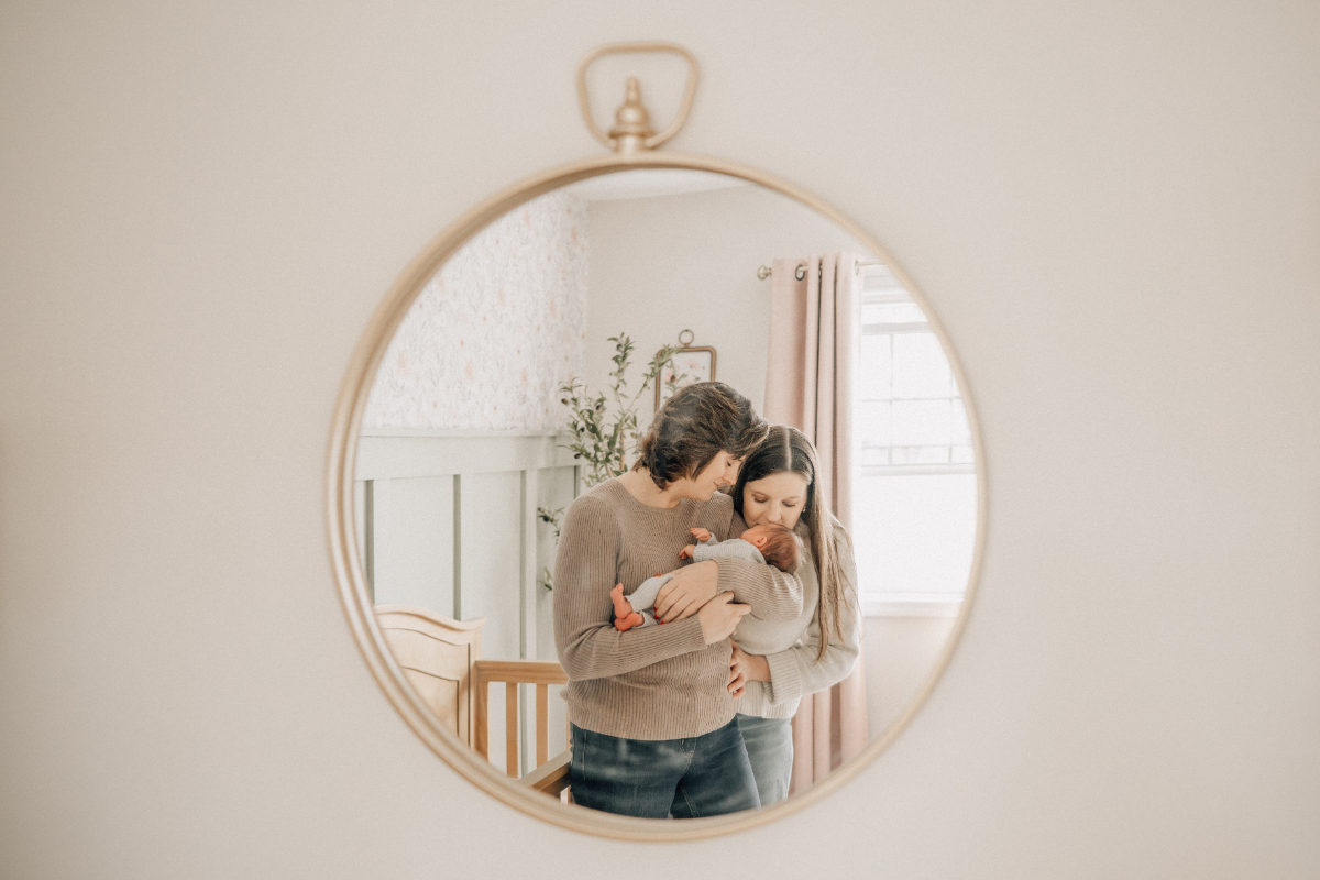 In-home newborn baby photos of two moms holding baby in nursery mirror.