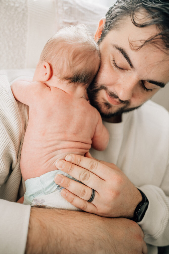 Father holding newborn against his chest, eyes closed in quiet, tender moment.