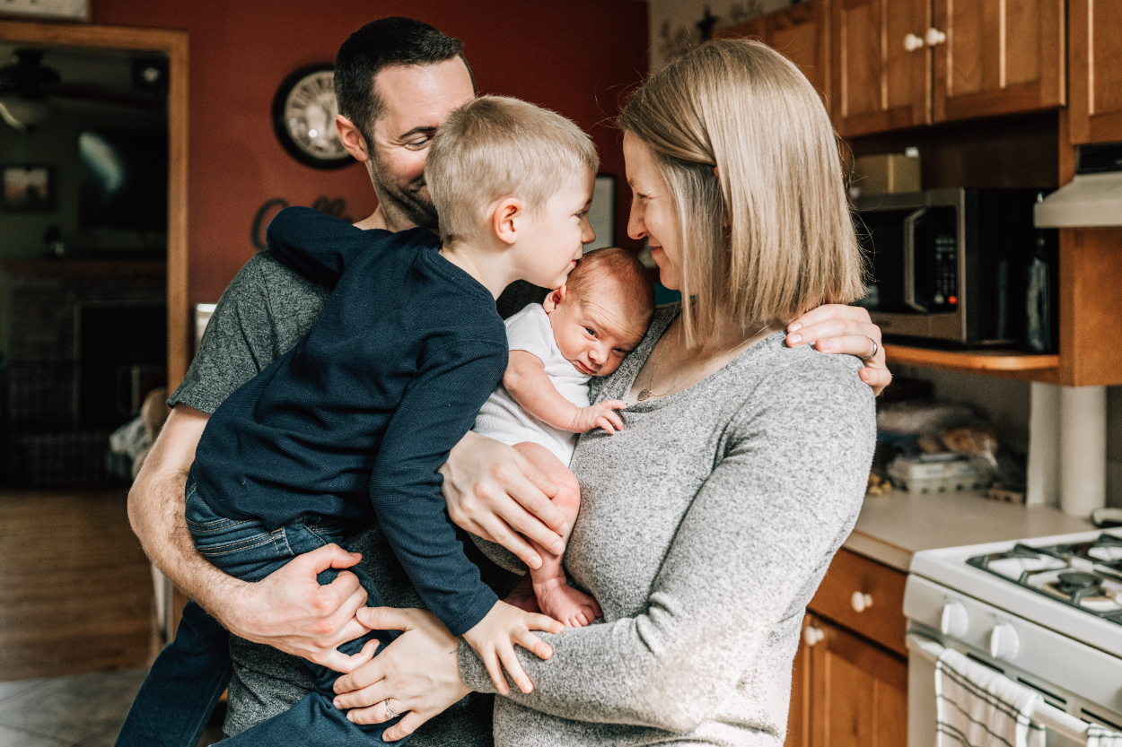 In-home newborn photography ideas with parents and sibling cuddling baby in kitchen.