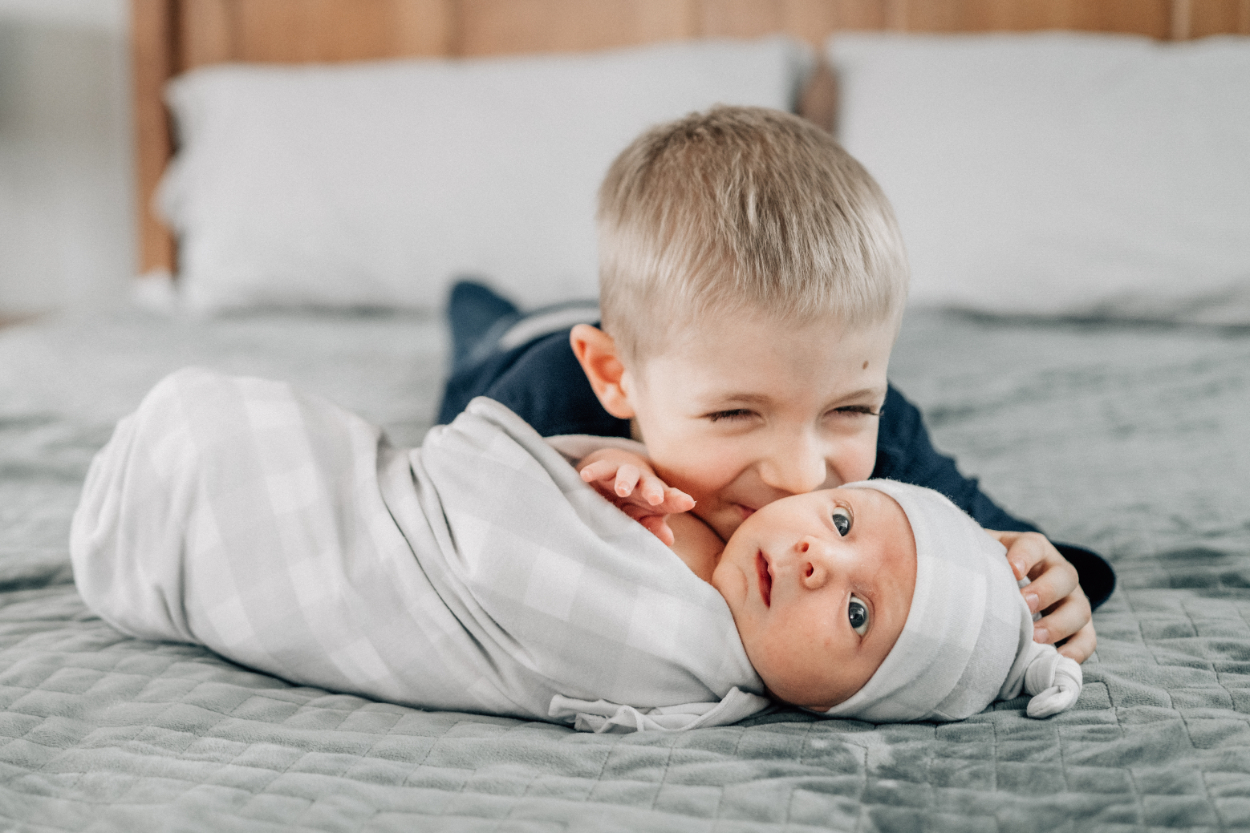 Newborn photography ideas with sibling gently hugging swaddled baby on bed.