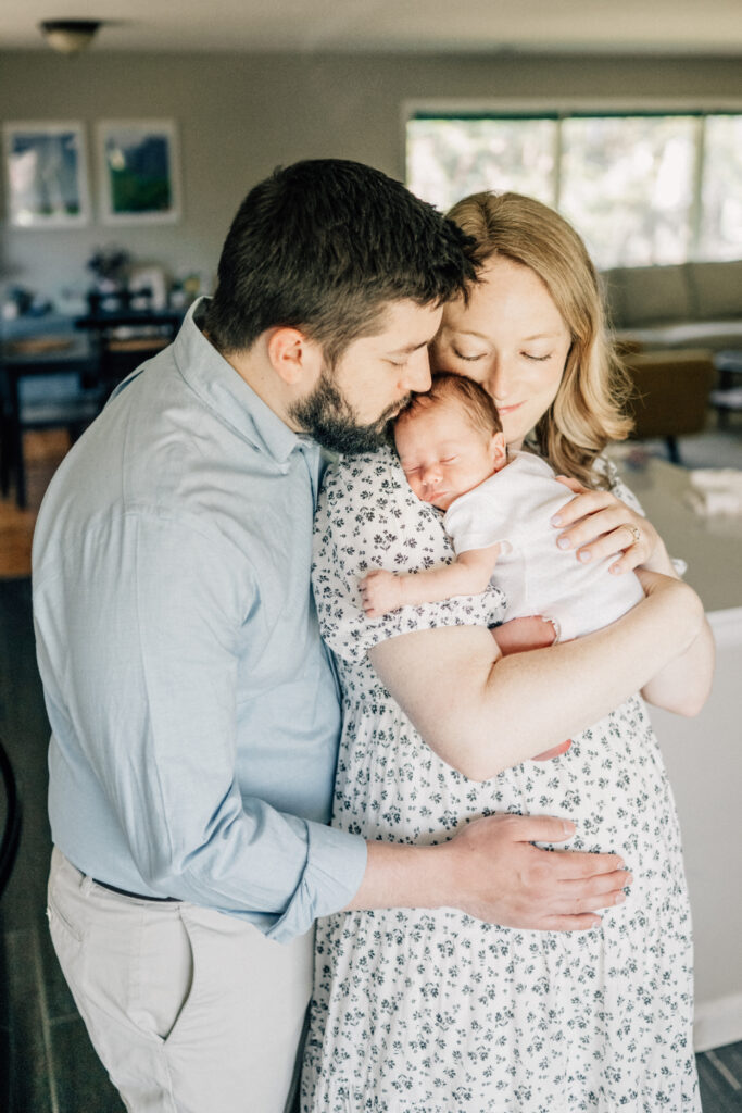 Parents holding newborn together, both kissing baby’s head in close family embrace.