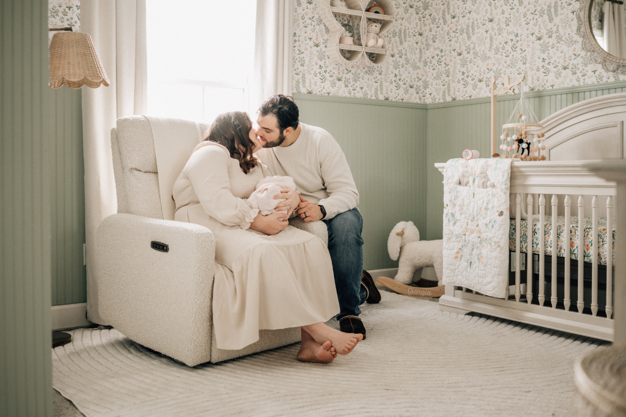 Parents sitting together in nursery chair, kissing baby while holding newborn close.