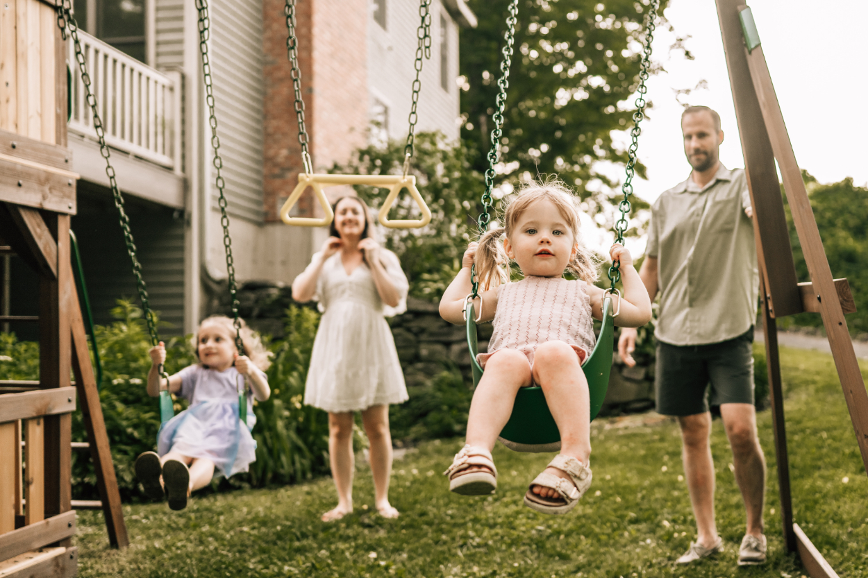 Kids on swings with parent nearby during a backyard photoshoot, showing playful movement and family connection
