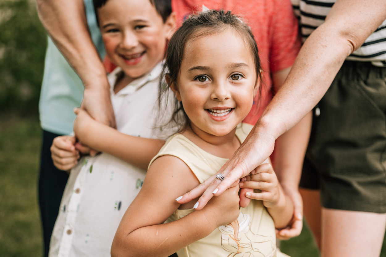 Child smiling while being hugged by parents during a backyard photoshoot, capturing warm family connection