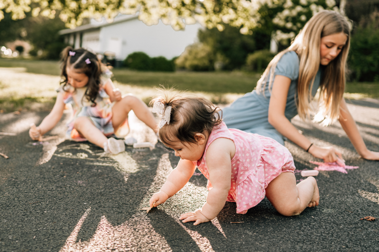 Children drawing with chalk on pavement while parent sits nearby, capturing playful and quiet outdoor moments