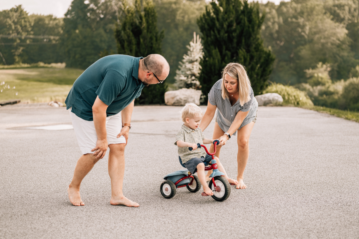 Parents helping toddler ride a tricycle in driveway, capturing supportive, everyday family moments outdoors