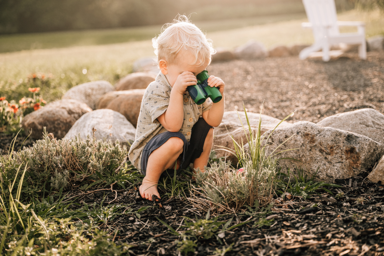 Toddler sitting near rocks looking through binoculars, capturing quiet, curious outdoor exploration moments