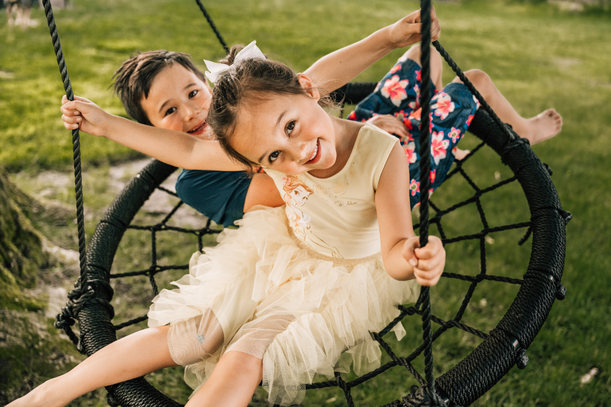 Two kids smiling on a swing during a backyard photoshoot, capturing playful, carefree childhood connection