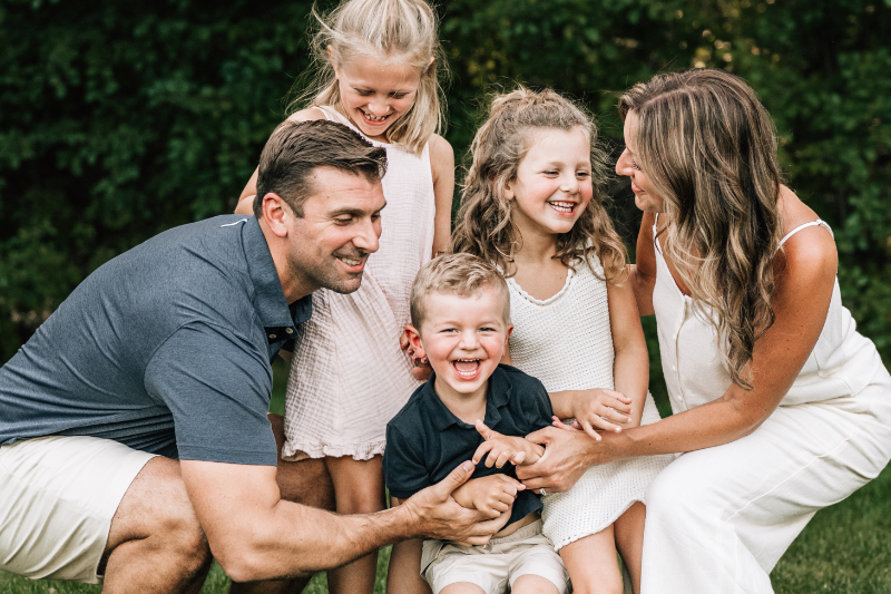 Family laughing together while parents playfully tickle their young son in the backyard.