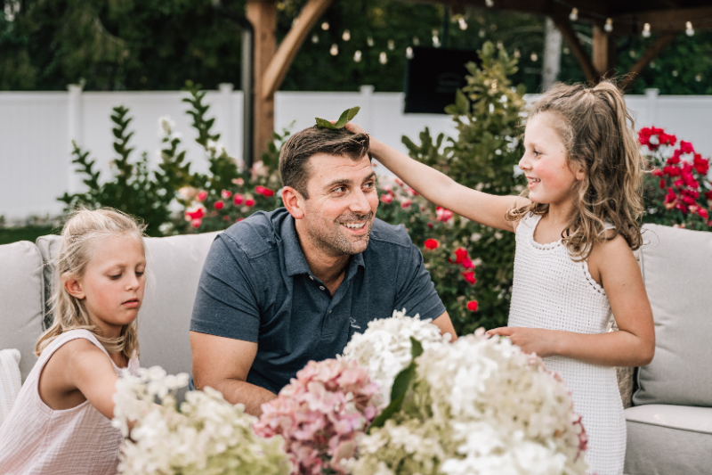 Child placing a leaf on father’s head while sitting together during playful outdoor family portraits in Albany NY backyard.