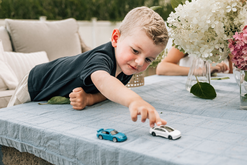 Child reaching across outdoor table to play with toy cars during backyard playtime.