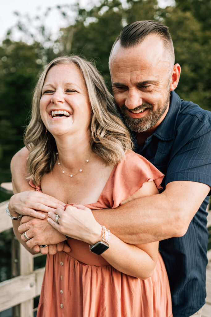 Couple embracing from behind, both smiling and laughing in warm natural light on wooden bridge