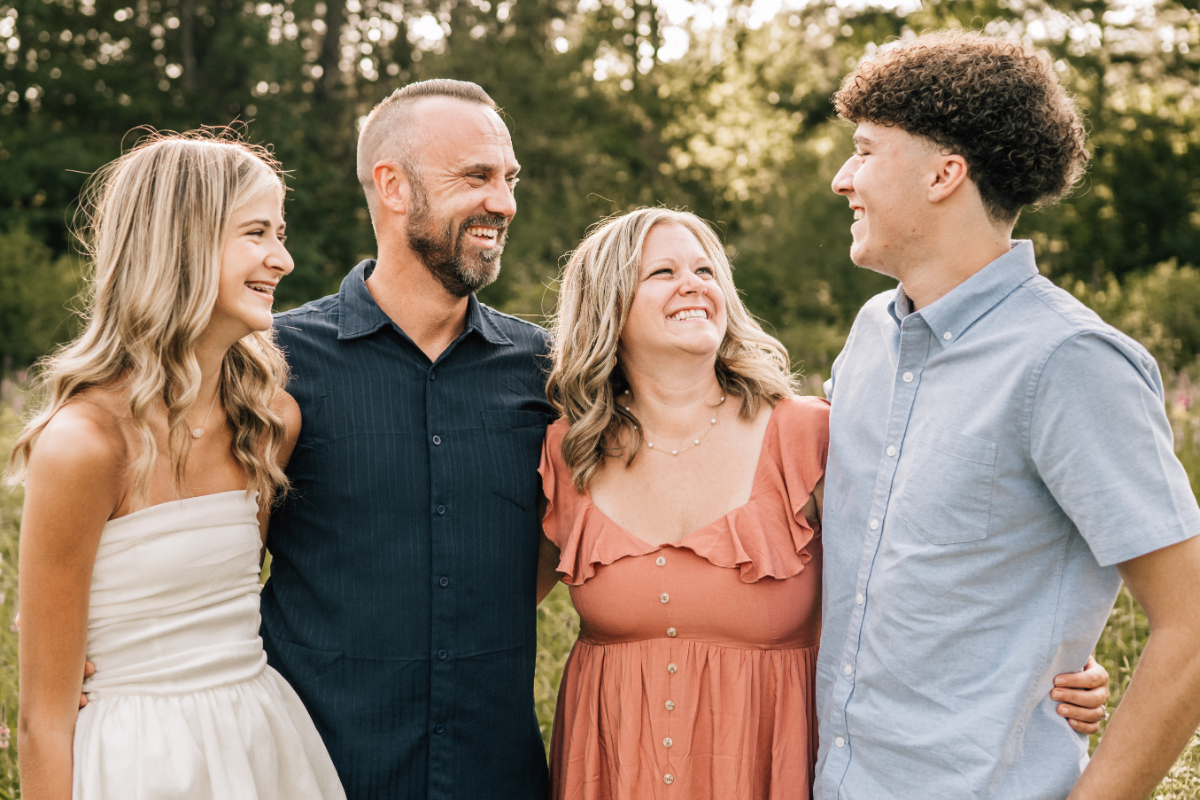 Family of four standing close together outdoors, smiling and laughing in warm natural light