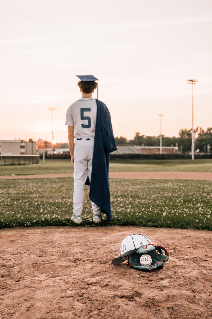 Baseball player standing on field at sunset wearing cap and uniform, holding jacket, graduation moment