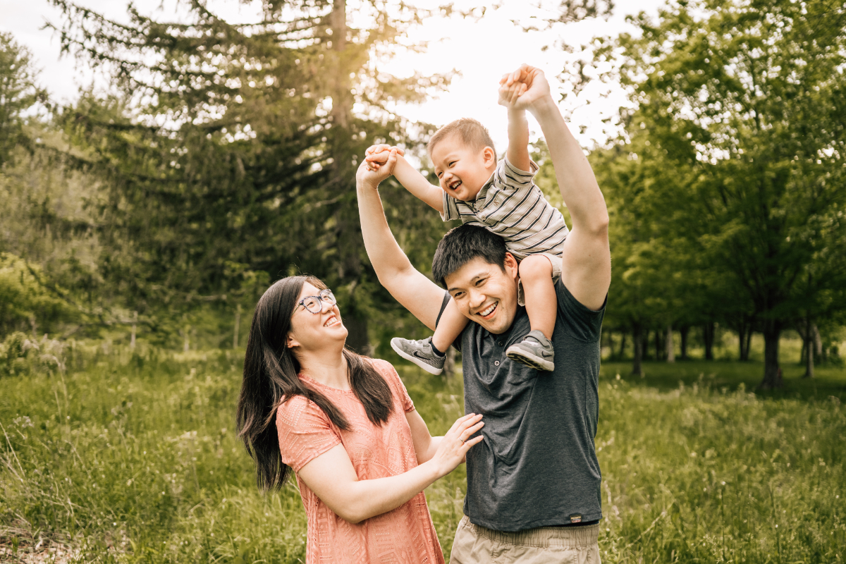 Spring family photos of happy parents lifting toddler on shoulders in soft green outdoor setting