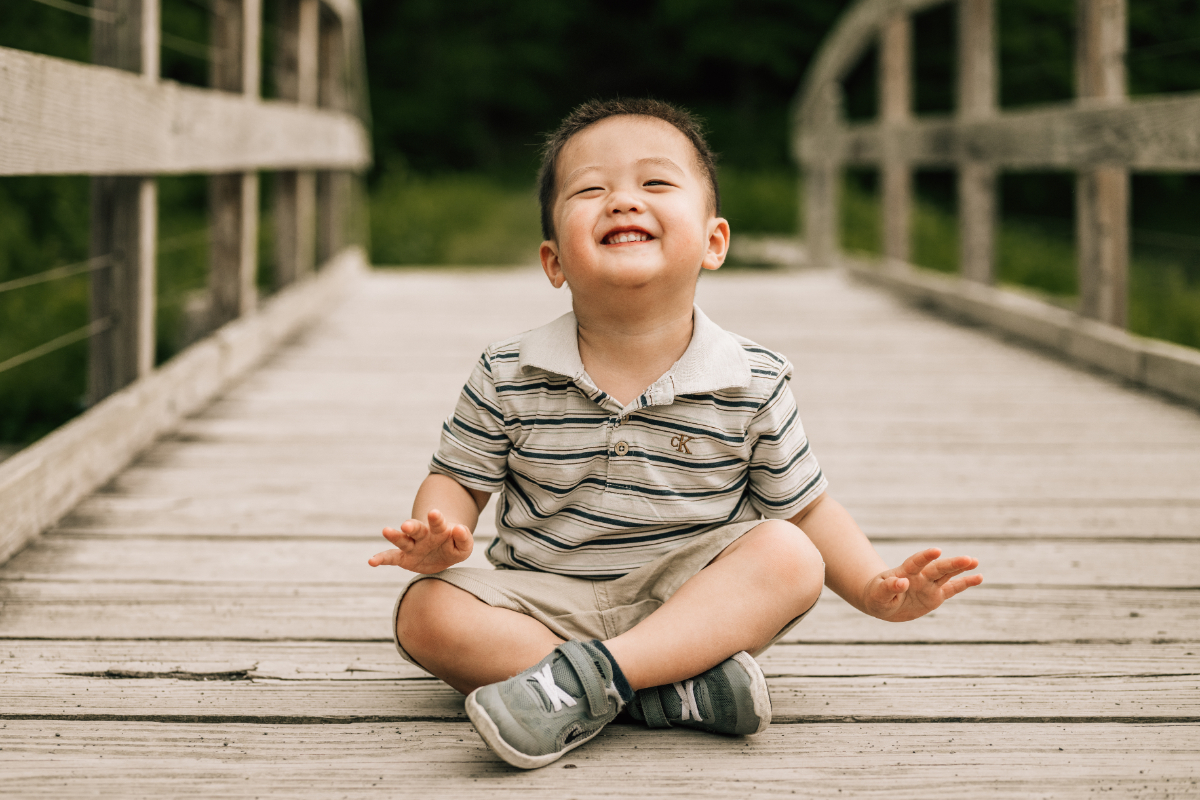 Smiling toddler sitting on wooden bridge, laughing with eyes closed in soft natural outdoor setting