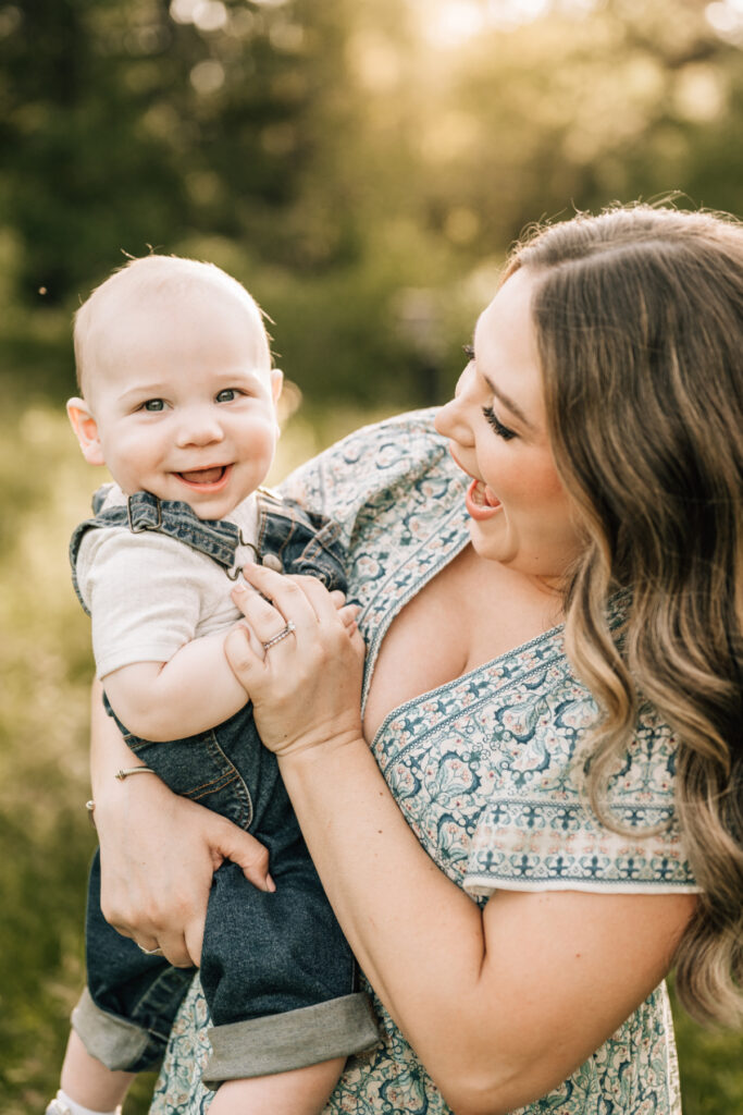 Spring family photos of mom holding smiling baby in warm sunlight, sharing candid, joyful moment outdoors