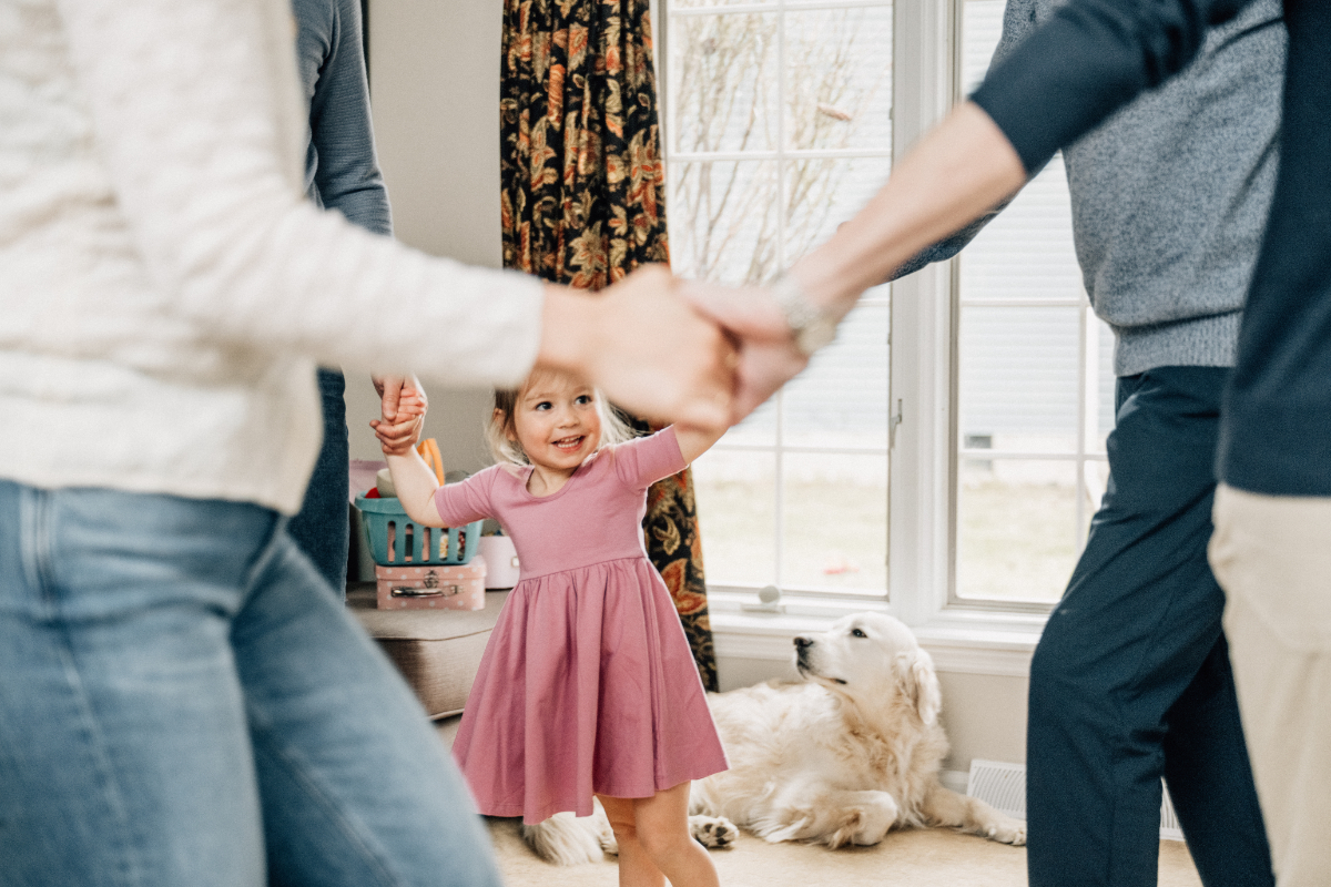 Parents holding child’s hands as she spins and laughs near window, joyful and active moment