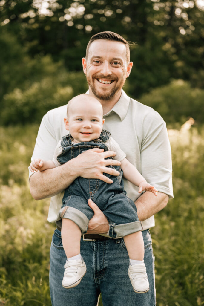 Spring family photos of dad holding baby in green field, both smiling in warm evening light