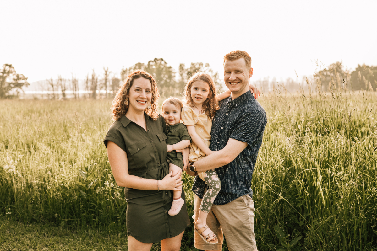 Family of four standing in tall grass field, smiling together in warm golden light outdoors