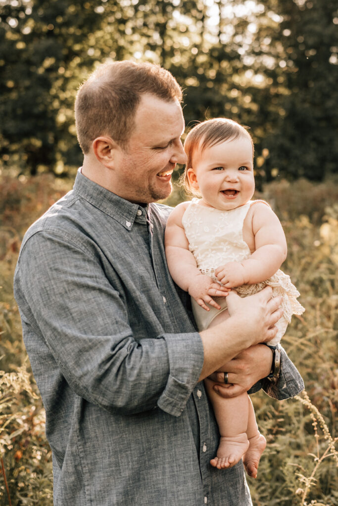 Dad holding smiling baby during baby’s first birthday outdoor session with warm golden light